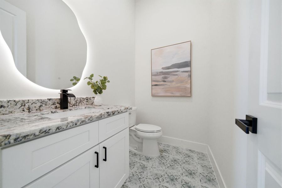 1st floor bathroom adjacent to the entry way. This is a modern, stylish bathroom featuring a sleek white vanity with a granite countertop, a large backlit mirror, and elegant patterned tile flooring.