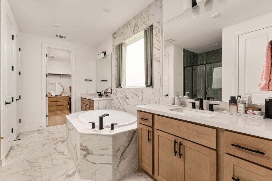 Freestanding soaking tub with marble-look surround, dual vanity with light wood cabinetry, white countertops, and black fixtures