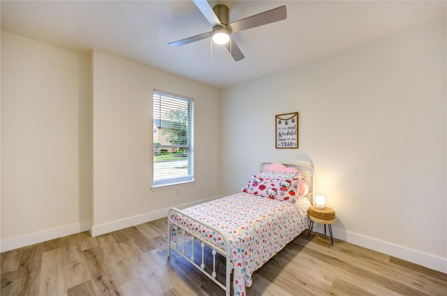 This cozy bedroom includes a ceiling fan, a large window providing ample natural light, The wood flooring creating a welcoming space.