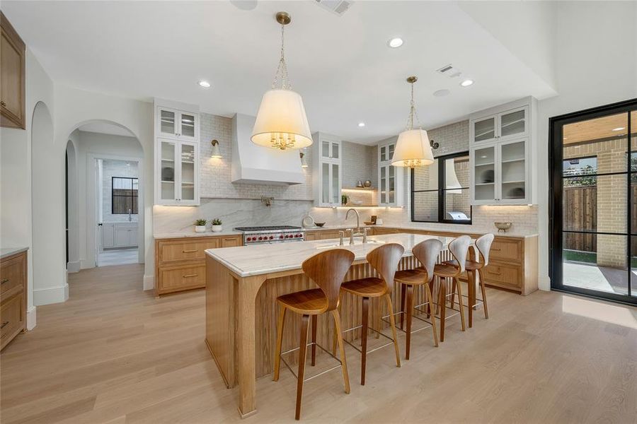 Gourmet kitchen with a large central island, light wood-finish flooring, white shaker-style upper cabinetry with glass inserts, recessed lighting, and a full-height tile backsplash
