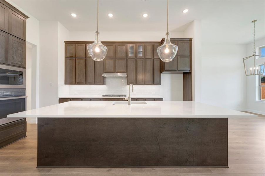 Spacious kitchen featuring a large central island with a white countertop, dark wood-finish cabinetry, built-in stainless steel appliances, and wood-finish flooring Spacious kitchen featuring a large central island with a white countertop, dark wood-finish cabinetry, built-in stainless steel appliances, and wood-finish flooring