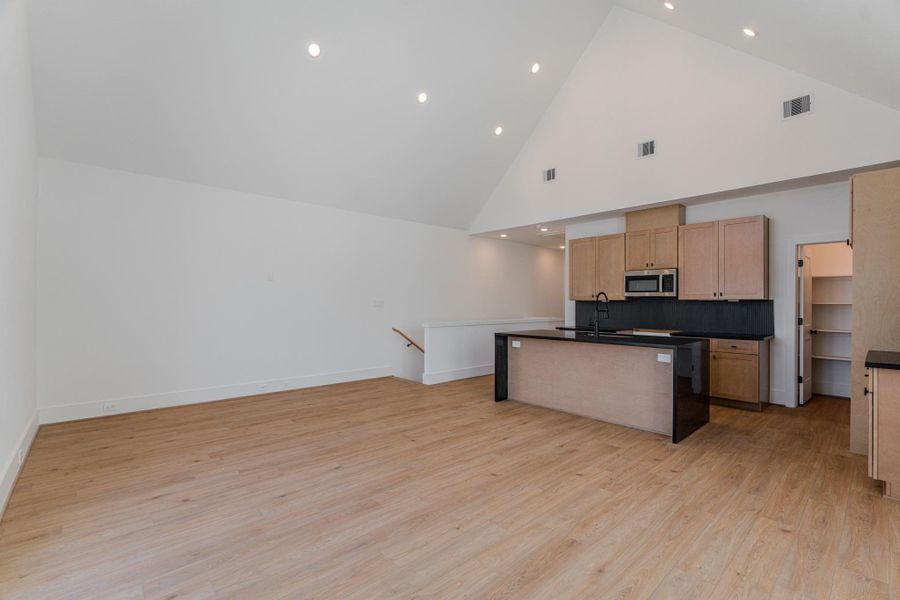 This photo showcases a spacious open-concept room with high vaulted ceilings and recessed lighting. It features a modern kitchen with light wood cabinets, a large central island, stainless steel appliances, and adjacent pantry storage. The room has light wood flooring and a clean, minimalist design.Photo Disclaimer: Photos are representative of the community and may depict a similar floor plan, model home, or previously completed residence. This photo showcases a spacious open-concept room with high vaulted ceilings and recessed lighting. It features a modern kitchen with light wood cabinets, a large central island, stainless steel appliances, and adjacent pantry storage. The room has light wood flooring and a clean, minimalist design.Photo Disclaimer: Photos are representative of the community and may depict a similar floor plan, model home, or previously completed residence.
