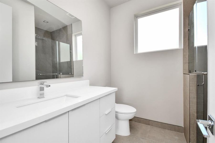 This modern bathroom features sleek white cabinetry, a large mirror, a walk-in shower with glass doors, and neutral tile finishing.