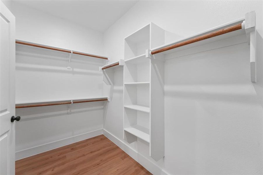 Walk-in closet featuring wood-finish flooring, white built-in shelving, two levels of hanging rods, and a white six-panel door with black hardware Walk-in closet featuring wood-finish flooring, white built-in shelving, two levels of hanging rods, and a white six-panel door with black hardware