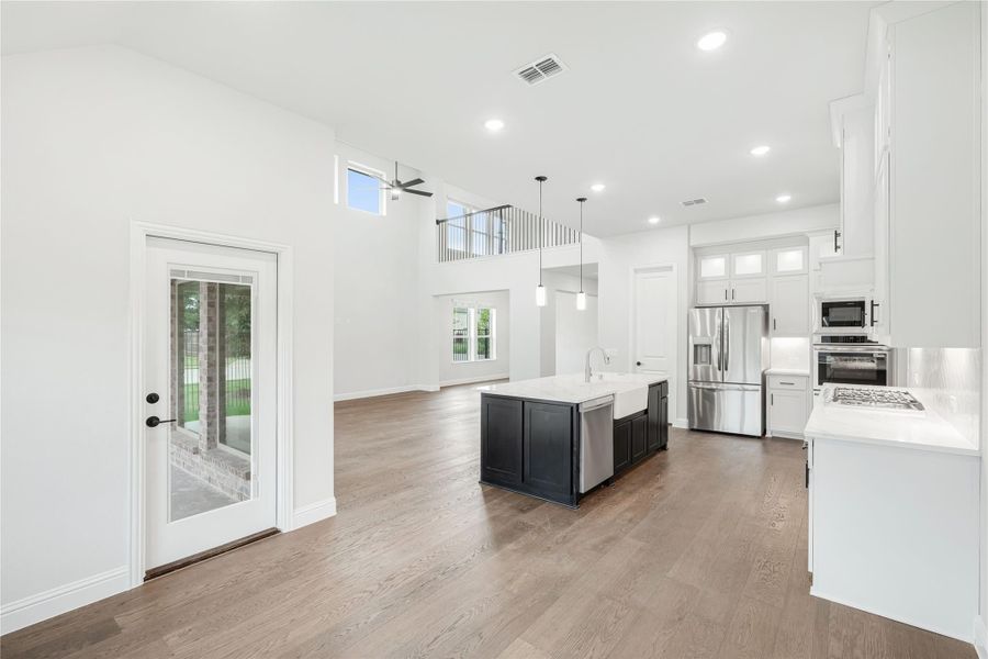 Open-concept kitchen with dark island, white cabinets, stainless appliances, and hardwood floors with two-story ceiling Open-concept kitchen with dark island, white cabinets, stainless appliances, and hardwood floors with two-story ceiling