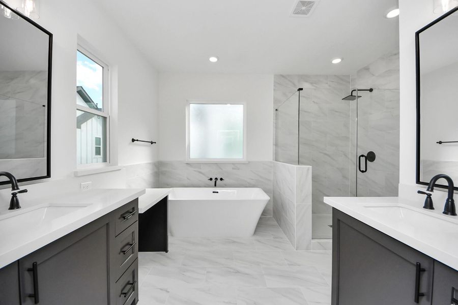 This bathroom is modern and spacious, featuring dual sinks with sleek black fixtures, a large soaking tub, and a glass-enclosed walk-in shower. The room is bright with ample natural light from the window, and the neutral tones create a serene atmosphere.