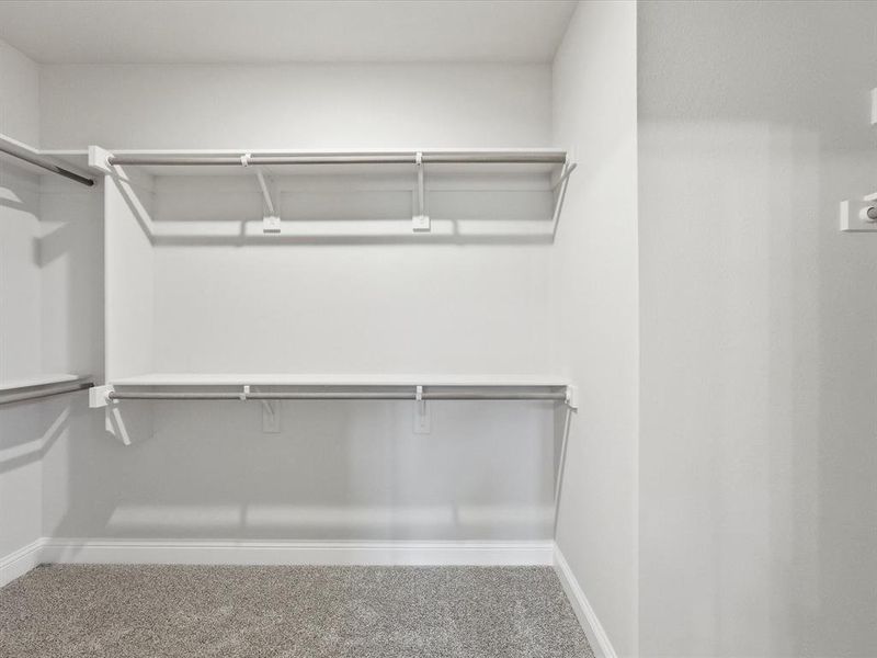 Spacious walk-in closet featuring white wire shelving and hanging rods, light gray wall paint, white baseboards, and gray carpet Spacious walk-in closet featuring white wire shelving and hanging rods, light gray wall paint, white baseboards, and gray carpet