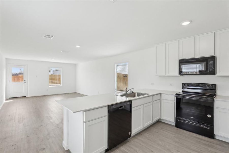 Kitchen featuring an island with a stainless steel sink, white shaker cabinetry, light-colored countertops, and black appliances