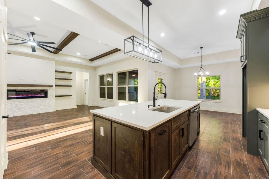 The modern open-concept living space features a spacious kitchen with a large island, contemporary light fixtures, and wood-look flooring. The living area includes a sleek electric fireplace and built-in shelves, complemented by large windows for natural light.