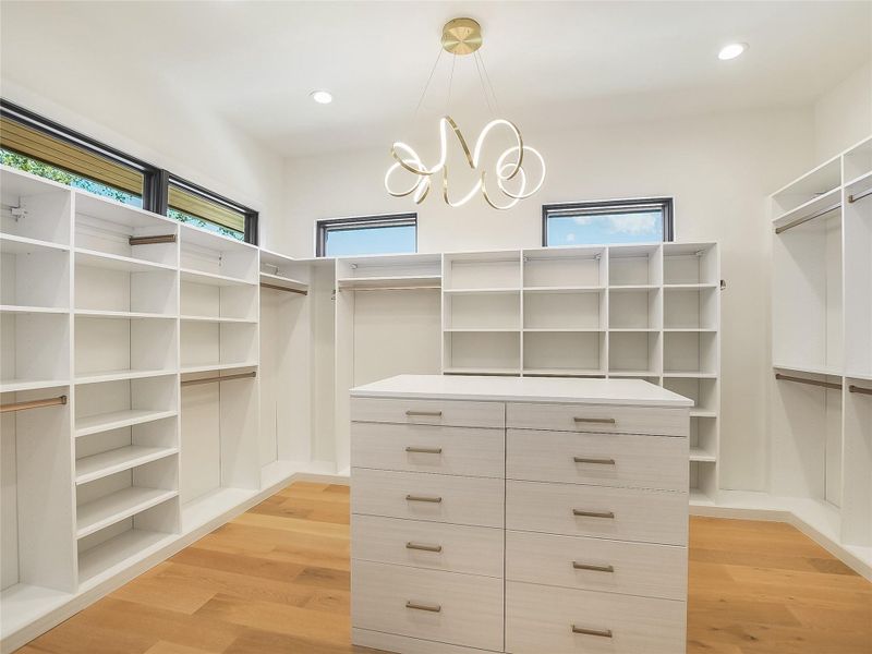 Spacious closet featuring extensive shelving, hanging rods, and a central island with drawers, all illuminated by recessed lighting and a contemporary chandelier