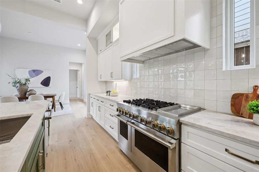 Two tone kitchen with light stone counters, range with two ovens, glass fronted cabinets, light wood finished floors, and backsplash