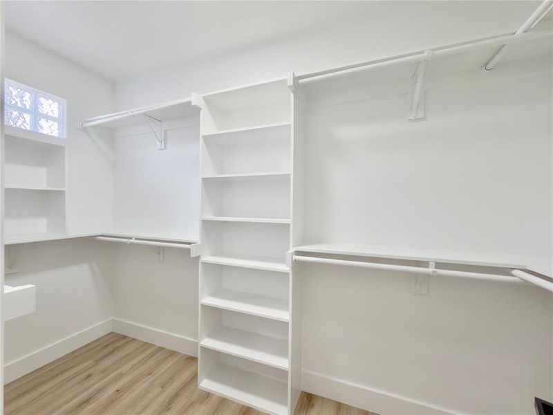 Spacious walk-in closet featuring light wood-finish flooring, white built-in shelving, white hanging rods, and a glass block window
