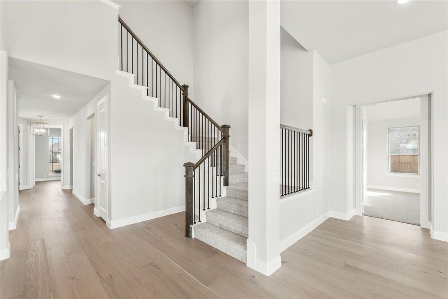 Bright foyer with carpeted staircase, dark iron railings, and light hardwood floors leading to hallway Bright foyer with carpeted staircase, dark iron railings, and light hardwood floors leading to hallway