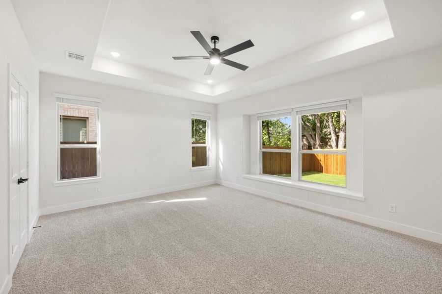 Empty bedroom with tray ceiling, ceiling fan, carpet flooring, and multiple windows with backyard views
