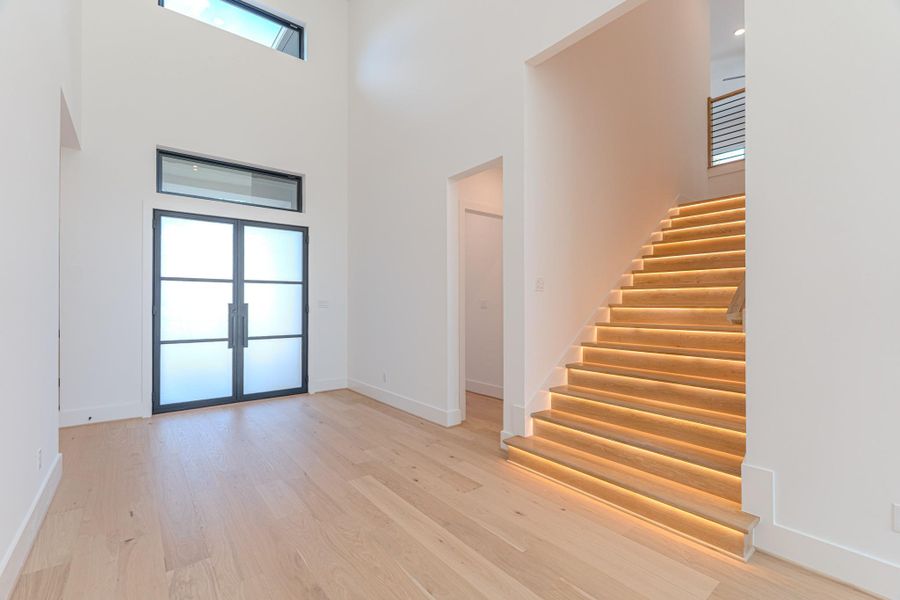 This photo showcases a modern entryway with high ceilings, large frosted glass doors, and sleek, illuminated wooden stairs. The space is bright and open, featuring light wood flooring and a minimalist design.