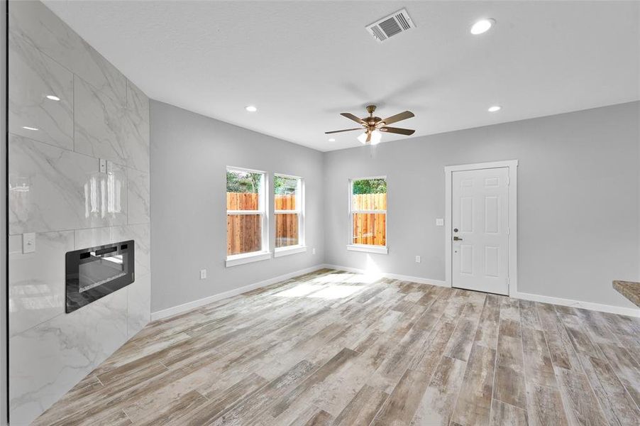 Step into a contemporary oasis with this sunlit living space, boasting an elegant wall-mounted electric fireplace, expansive windows, stylish wood-inspired flooring, a modern ceiling fan, and a soothing neutral palette.