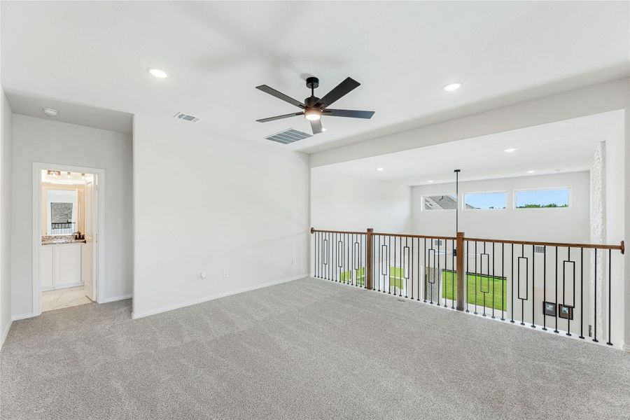 Open upstairs loft with gray carpet, ceiling fan, metal stair railing, and view into bathroom