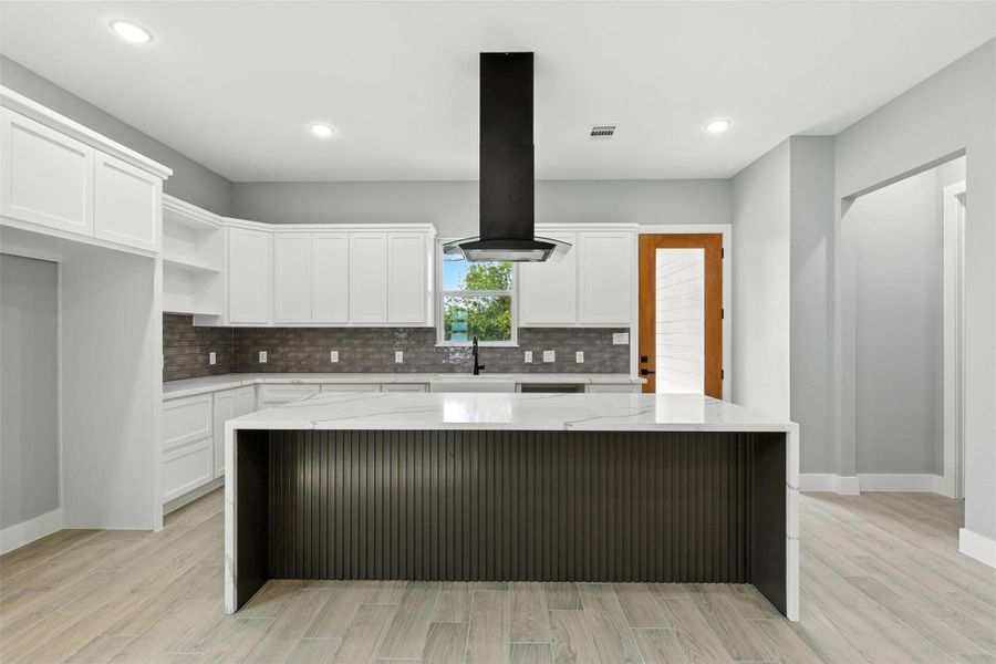 This modern kitchen features sleek white cabinetry, a central island with a black range hood, and a stylish backsplash. The space is bright and open, with a mix of wood and tile flooring, creating a clean and inviting atmosphere.