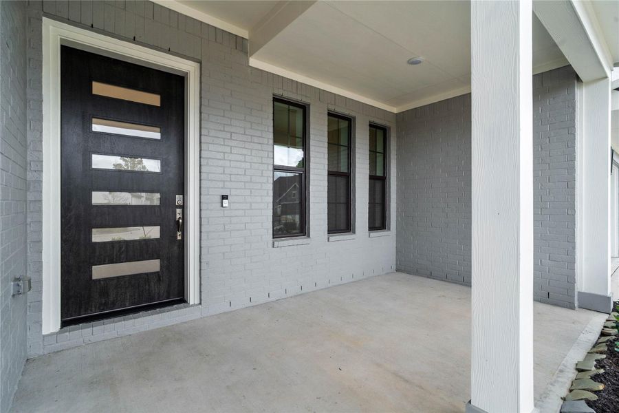 This covered front porch offers a clean, contemporary feel with its sleek grey brick façade and smooth concrete flooring. The space is anchored by a stunning modern five-lite glass front door in a rich dark finish, which provides an inviting and sophisticated entrance to the home.