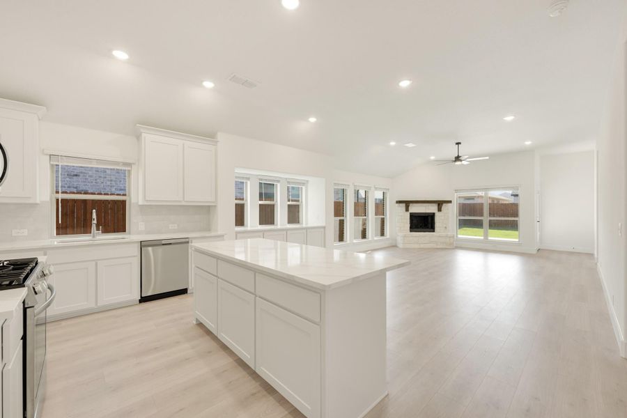 Open-concept kitchen with white cabinets and island flowing into living room with stone fireplace and light wood floors Open-concept kitchen with white cabinets and island flowing into living room with stone fireplace and light wood floors