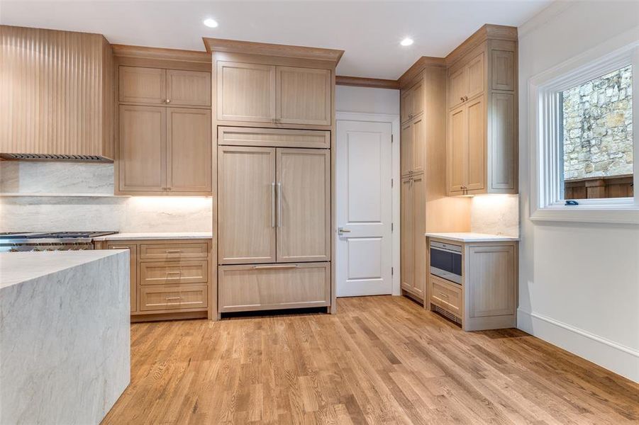 Kitchen featuring tasteful backsplash, paneled refrigerator, light wood-style floors, light stone countertops, and wall chimney exhaust hood Kitchen featuring tasteful backsplash, paneled refrigerator, light wood-style floors, light stone countertops, and wall chimney exhaust hood