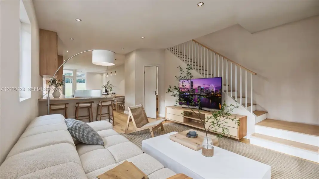 Open-concept interior featuring light wood-finish flooring, recessed lighting, a kitchen breakfast bar, and a staircase with white vertical balusters and a wood-finish handrail Open-concept interior featuring light wood-finish flooring, recessed lighting, a kitchen breakfast bar, and a staircase with white vertical balusters and a wood-finish handrail