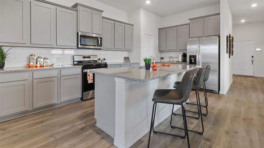 Kitchen with gray cabinetry, stainless steel appliances, a breakfast bar, an island with sink, and light stone counters