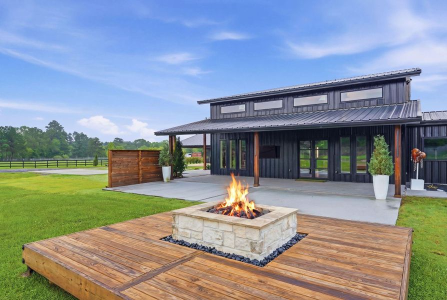 Modern home with a sleek black exterior and large windows. The backyard features a spacious patio with a built-in fire pit on a wooden deck, surrounded by lush green lawn and scenic views.