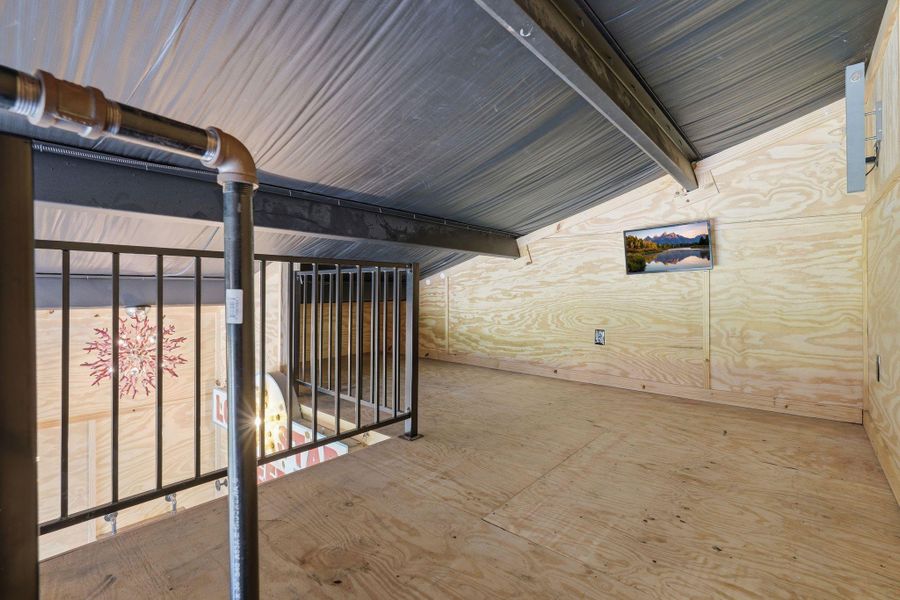 This photo shows a compact loft space with plywood walls and flooring, featuring a metal railing overlooking the area below. The ceiling is angled, giving a cozy feel over the home gym.