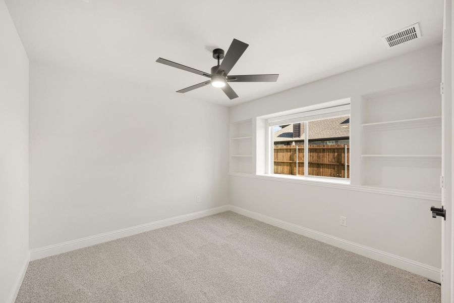 Empty bedroom with carpet, ceiling fan, built-in shelves, and a window overlooking a backyard fence