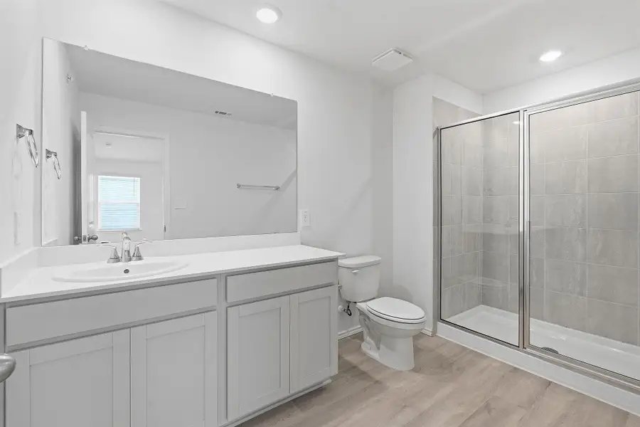 Bathroom featuring a spacious vanity with white cabinetry and an undermount sink, a large frameless mirror, a commode, a glass-enclosed shower with tiled walls, and wood-finish flooring Bathroom featuring a spacious vanity with white cabinetry and an undermount sink, a large frameless mirror, a commode, a glass-enclosed shower with tiled walls, and wood-finish flooring