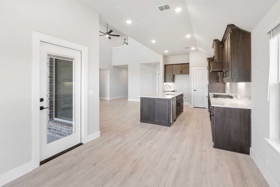 Open kitchen with dark wood cabinets, center island, light hardwood floors, and glass back door leading to patio. Open kitchen with dark wood cabinets, center island, light hardwood floors, and glass back door leading to patio.