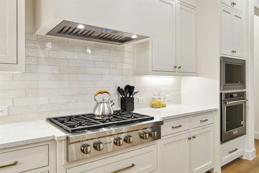 Kitchen featuring appliances with stainless steel finishes, backsplash, light stone counters, white cabinets, and wall chimney exhaust hood