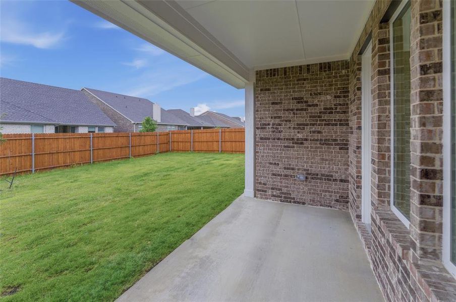 Covered patio with a concrete surface, brown brick exterior, and large windows overlooking a spacious, fenced backyard with manicured lawn