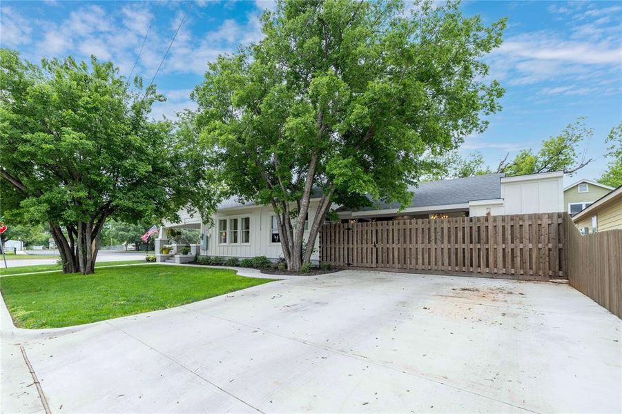 White exterior residence featuring a dark shingled roof, a concrete driveway, a wooden privacy fence, and mature trees