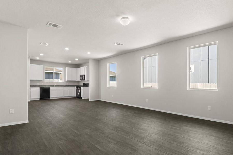 Image of home living areas with kitchen in rear with windows on one wall and over kitchen sink. There are wood-look floors and white walls