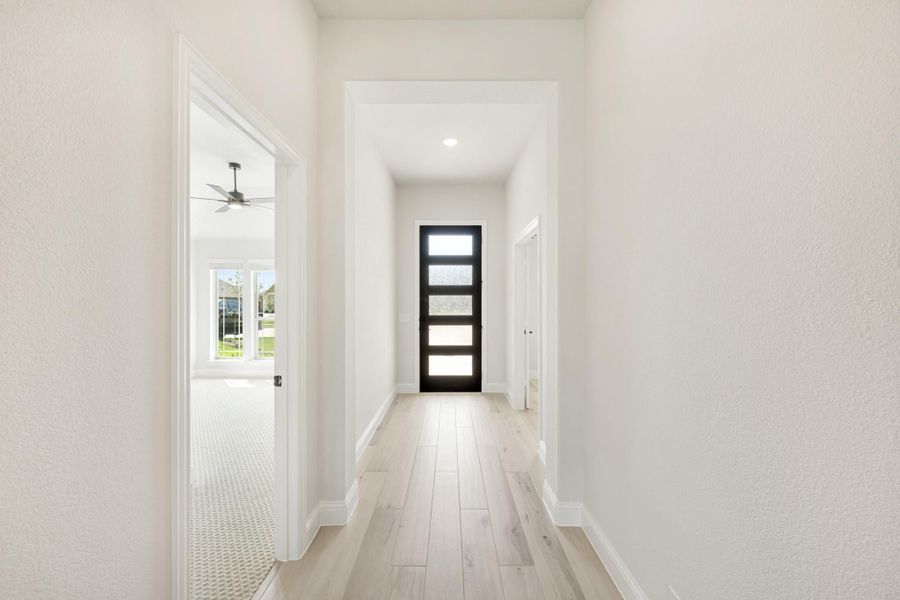 Hallway with light wood floors, white walls, and a black glass-panel door at the end