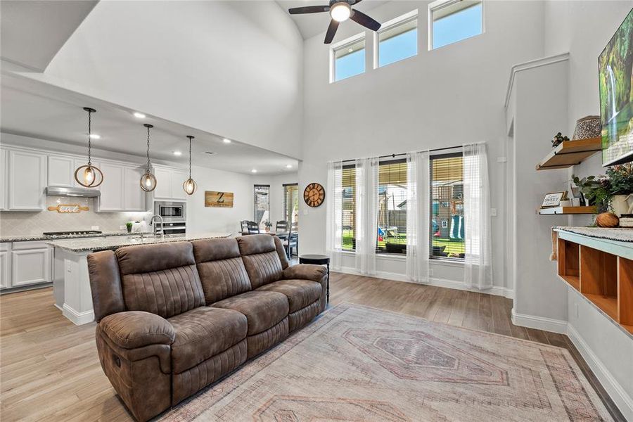 This is an open-plan living space with vaulted ceilings and large windows providing plenty of natural light. The area features a modern kitchen a cozy living area and stylish pendant lighting above the kitchen island. The room has a neutral color palette and wood flooring, creating a warm and inviting atmosphere.