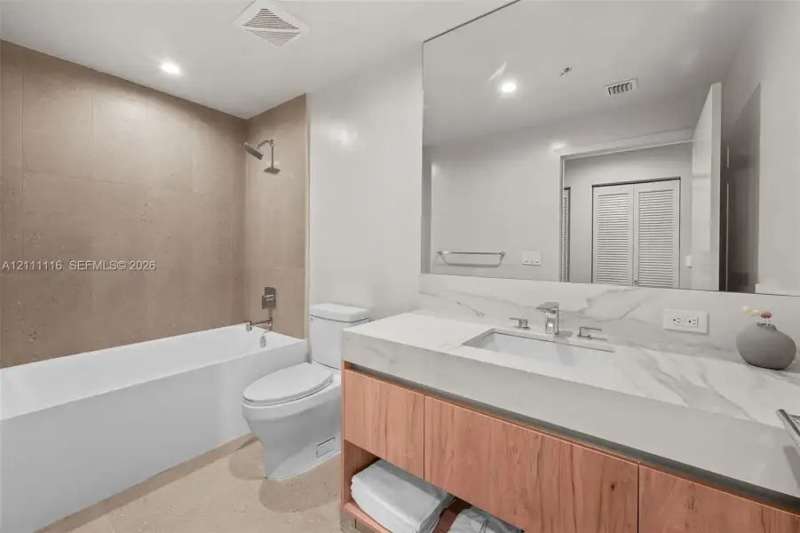 Bathroom featuring a wood-finish floating vanity with stone-surround countertop, a large wall-mounted mirror, a deep soaking tub with a floor-to-ceiling neutral tile surround, and recessed ceiling lighting