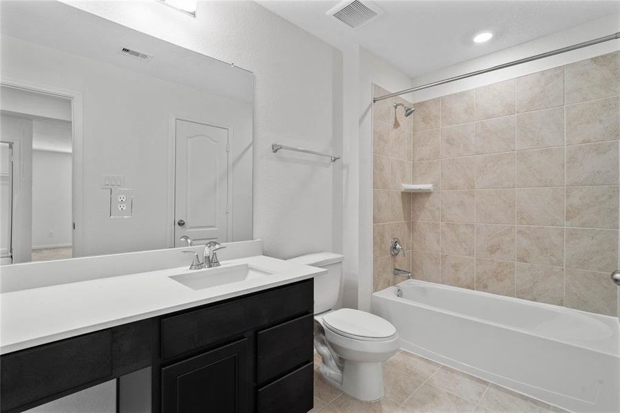 Secondary bath features tile flooring, bath/shower combo with tile surround, stained wood cabinets, beautiful light countertops, mirror, dark, sleek fixtures and modern finishes.