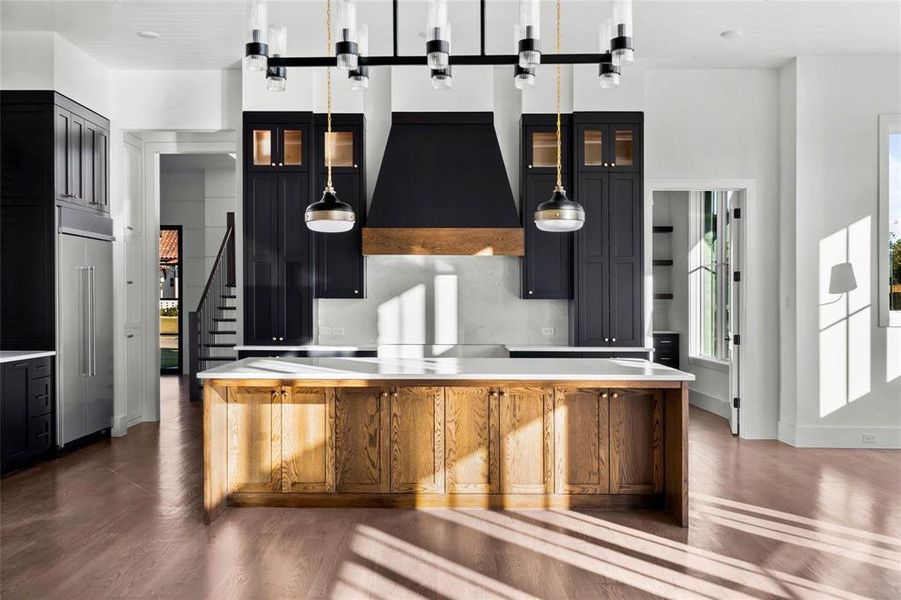Kitchen with dark cabinets, glass insert cabinets, wall chimney exhaust hood, and a spacious island Kitchen with dark cabinets, glass insert cabinets, wall chimney exhaust hood, and a spacious island