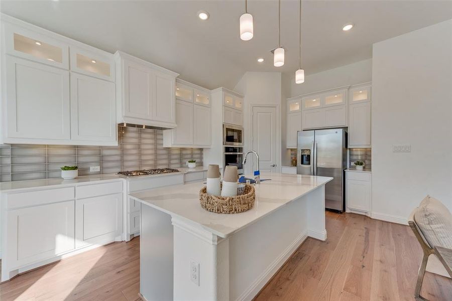 Kitchen featuring a center island with sink, stainless steel appliances, hanging light fixtures, decorative backsplash, and white cabinets