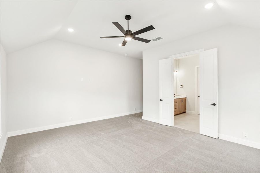 Empty bedroom with vaulted ceiling, ceiling fan, gray carpet, and open door to ensuite bathroom