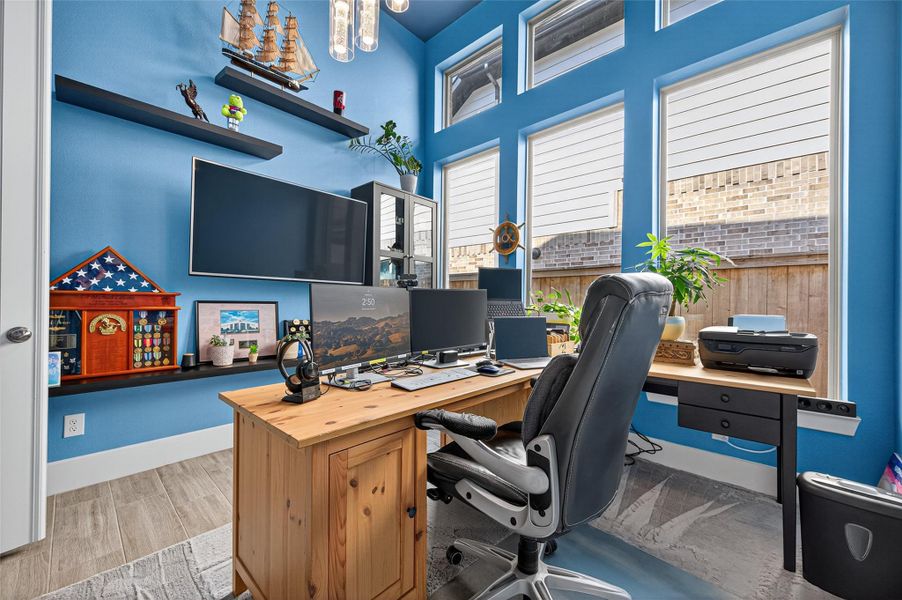 This is a well-lit home office with blue walls and large windows. It features a wooden desk with multiple monitors, a comfortable black office chair, and a variety of decor including shelves, plants, and nautical-themed items. The room is organized and inviting, ideal for productivity. This is a well-lit home office with blue walls and large windows. It features a wooden desk with multiple monitors, a comfortable black office chair, and a variety of decor including shelves, plants, and nautical-themed items. The room is organized and inviting, ideal for productivity.
