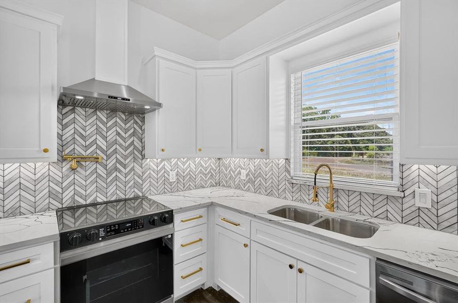 Kitchen featuring white cabinetry with gold hardware, a herringbone pattern tile backsplash, light-toned countertops, and a stainless steel appliance suite including a range and hood