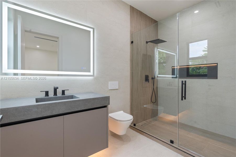 Floating vanity with integrated sink and matte black faucet, wall-mounted toilet, walk-in shower with glass enclosure, wood-finish tile shower wall, and illuminated vanity mirror