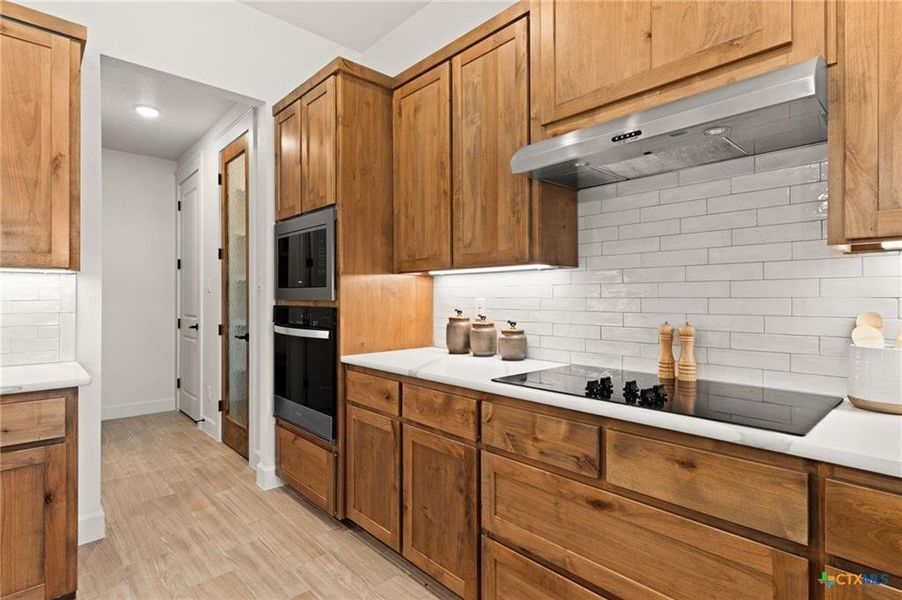 Kitchen with tasteful backsplash, brown cabinets, range hood, black appliances, and light wood-style floors