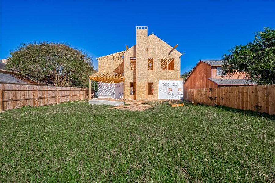 This photo showcases a partially constructed two-story home with wooden framing and exterior sheathing. It features a spacious fenced backyard with green grass, surrounded by neighboring homes and trees.