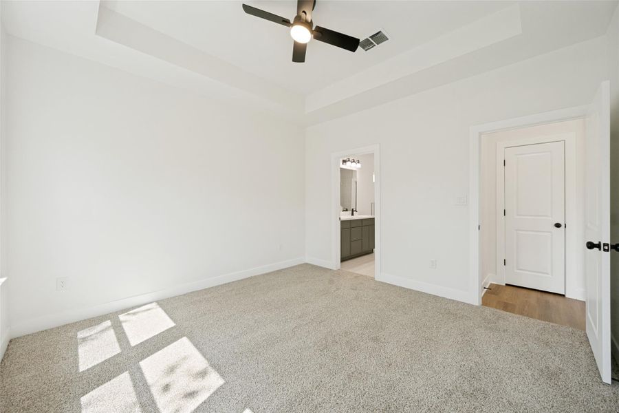 A spacious primary bedroom features soft carpeting, a tray ceiling, and a ceiling fan, with a private bathroom visible through an open doorway. A spacious primary bedroom features soft carpeting, a tray ceiling, and a ceiling fan, with a private bathroom visible through an open doorway.