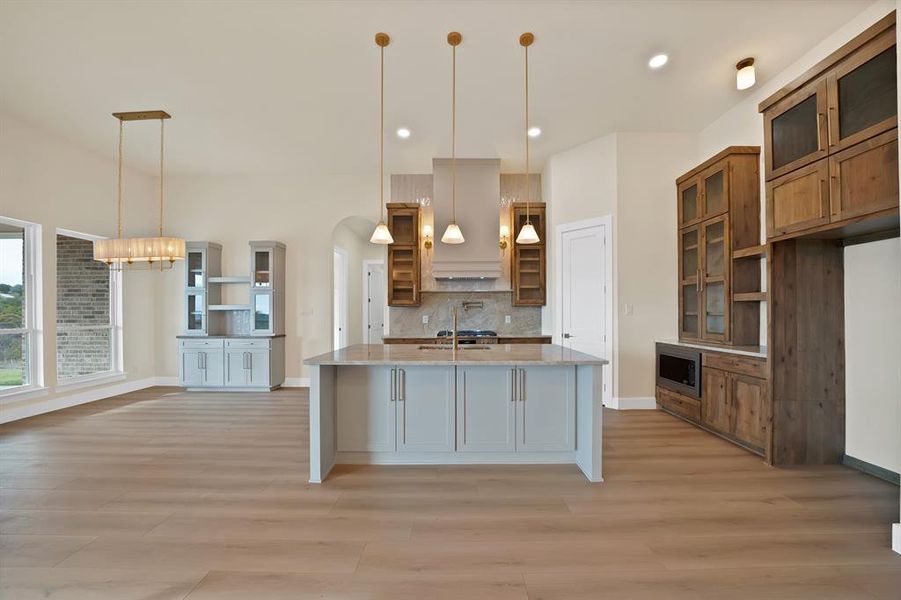 Kitchen featuring glass insert cabinets, a kitchen island with sink, pendant lighting, light stone countertops, and decorative backsplash Kitchen featuring glass insert cabinets, a kitchen island with sink, pendant lighting, light stone countertops, and decorative backsplash